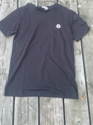 Svart Moncler t-shirt med logga - Snygg svart t-shirt från Moncler med klassisk rund hals och korta ärmar. På bröstet sitter Monclers ikoniska broderade logga i vitt, blått och rött. T-shirten är i mjuk bomull och har en clean, stilren look.