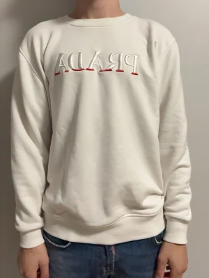Vit sweatshirt från Prada - Vit sweatshirt från Prada med broderad logga i vitt och rött framtill. Klassisk rund halsringning och ribbade muddar vid ärmslut och nederkant. Perfekt för en clean och stilren look.