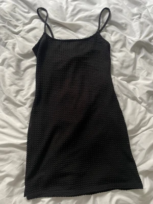 Svart kort klänning med struktur - Säljer en svart kort klänning med smala axelband och snygg strukturerad yta. Klänningen har en figurnära passform och är perfekt för dig som gillar enkel men stilren design. Materialet känns mjukt och stretchigt, vilket gör den bekväm att bära.