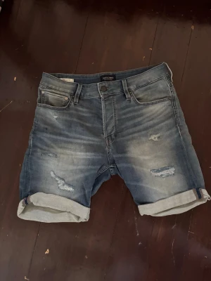 Jeansshorts från Jack & Jones - Blå jeansshorts från Jack & Jones med slitna detaljer och uppvikta benslut. Klassisk femficksmodell med knappgylf och bälteshällor. 