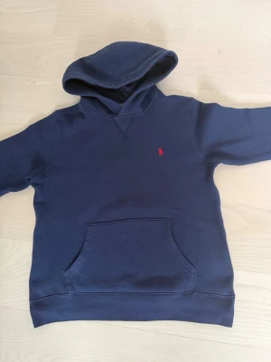 Mörkblå hoodie från Ralph Lauren - Mörkblå hoodie från Ralph Lauren med klassisk röd logga broderad på bröstet. Hoodien har en stor känguruficka framtill och en skön huva. Tillverkad i mjukt bomullsmaterial som känns bekvämt mot huden. Perfekt för en avslappnad och stilren look. Pris kan diskuteras 
