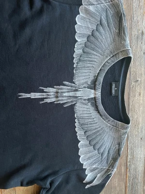 Svart Hoodie med vingeprint från Marcelo Burlon - Säljer en svart Hoodie från Marcelo Burlon med ikoniskt grått vingeprint över axlar och bröst, samt på ryggen. Hoodien har rund hals och armar passande. Materialet är mjuk bomull och passformen är normal. Perfekt för dig som vill sticka ut med en unik design.