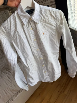 Randig skjorta från Ralph Lauren - Klassisk vit skjorta med tunna blå ränder från Ralph Lauren. Skjortan har button-down krage, långa ärmar och den ikoniska broderade loggan på bröstet. Tillverkad i bomull för en skön och luftig känsla. Perfekt till jeans eller chinos.