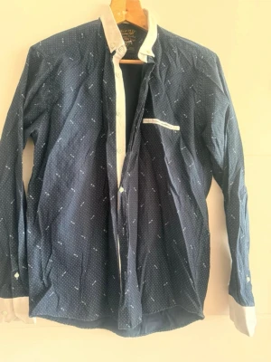 👔 Herrskjorta – marinblå med vita prickar - 👔 Herrskjorta – marinblå med vita prickar  Säljer en elegant herrskjorta 👕 i marinblå färg med ett fint mönster av små vita prickar ⚪️⚪️⚪️  Skjortan ser väldigt snygg ut ✨ och passar både till vardags 🏙️ och mer formella tillfällen 🎩  📏 Storlek: L 📐 Bröstvidd (under armarna): 100 cm 📏 Längd: 66 cm  🧵 Material:  * 60% bomull * 40% polyester  Materialet är bekvämt 👍, skönt att bära och sitter bra på kroppen 👌  Skick: mycket bra ⭐️ 
