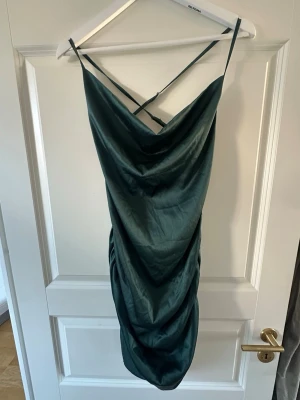 Mörkgrön satin klänning med smala band - Snygg mörkgrön klänning med smala axelband som korsas i ryggen. Klänningen har draperad halsringning och en figurnära passform som ger en elegant form. Den är ihopsydd under rumpan men inget som syns när den sitter på. Köpt secoundhand men endast använd en gång av mig! 