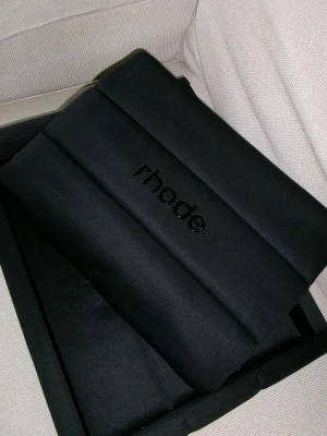 Rhode bubble bag in black  - Säljer en svart vadderad clutch från Rhode med stilren design och diskret logga framtill. Väskan har en mjuk, puffig form och är tillverkad i ett tygmaterial som ger en modern och trendig look. Perfekt accessoar för dig som gillar minimalistisk stil.
