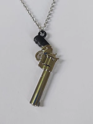 Halsband med pistolhänge i metall - Snyggt halsband med ett detaljerat pistolformat revolver hänge i silverfärgad metall och svart handtag. Kedjan är tunn och silvrig, vilket ger ett coolt och edgy intryck. Perfekt accessoar för dig som vill sticka ut och addera lite attityd till din stil. Materialet rostfritt stål 