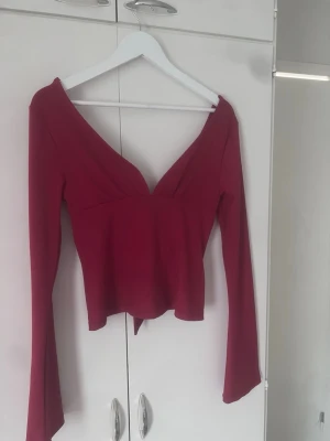 Mörkröd långärmad topp med knyt - Trendig mörkröd topp med lång ärm och djup v-ringning. Toppen har snygg knytning i ryggen som ger en unik detalj. Perfekt för dig som vill ha en stilren men ändå iögonfallande look.