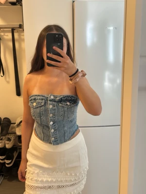 Blå jeans topp - Säljer en snygg blå tubtopp i jeans med silverfärgade knappar framtill och två bröstfickor. Toppen är axelbandslös och har en croppad passform.