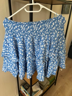 Blå volangkjol från SAMSA Collection - Supersöt blå kortkjol från SAMSA Collection med vitt blommigt mönster och bred smock i midjan. Kjolen har volanger som ger en flowy känsla och passar perfekt till sommaren. Tillverkad i ett lätt och skönt material.