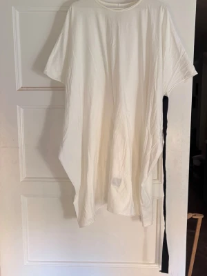 Vit oversized t-shirt från MM6 Maison Margiela - Vit oversized t-shirt från MM6 Maison Margiela och Rick Owens med asymmetrisk form och kort ärm. T-shirten har en unik svart detalj längs ena sidan och är tillverkad i mjuk bomull. Perfekt för en avslappnad och trendig look.