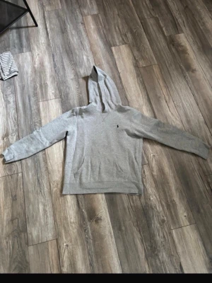 Grå hoodie från Polo Ralph Lauren - Klassisk grå hoodie från Polo Ralph Lauren med broderad logga på bröstet. Hoodien har huva, känguruficka och ribbade muddar vid ärmslut och nederkant. Tillverkad i mjukt bomullsmaterial som är skönt att ha på sig. Storleken är XL i barnstorlek som motsvarar XS eller S