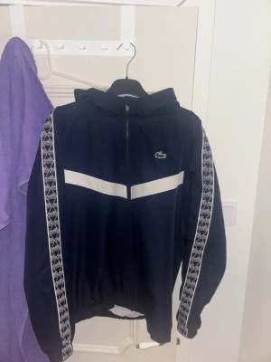 Marinblå vindjacka från Lacoste - Snygg marinblå vindjacka från Lacoste med vit rand över bröstet och mönstrade band längs ärmarna. Jackan har huva, dragkedja framtill och elastisk nederkant. Perfekt för dig som vill ha en sportig och stilren look.