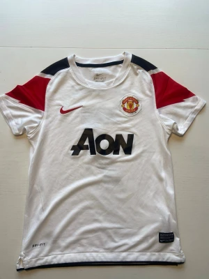 Manchester United bortatröja Nike - Snygg vit Manchester United bortatröja från Nike med röda och svarta detaljer. Tröjan har klubbmärke, sponsorlogga och nummer 7 med Di Maria på ryggen. Tillverkad i Dri-FIT material som andas, perfekt för fotboll eller träning.