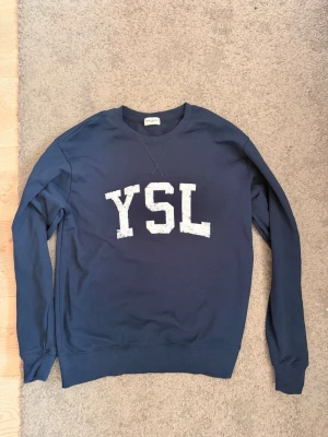 Mörkblå YSL sweatshirt Saint Laurent - Mörkblå sweatshirt från Saint Laurent med stort vitt YSL-tryck på bröstet. Klassisk rund halsringning och ribbade muddar vid ärmslut och nederkant. Tillverkad i mjukt bomullsmaterial för en skön och avslappnad look.