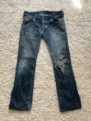 Blå slitna Dieseljeans med hål - Säljer ett par blå Dieseljeans med slitningar och ett stort hål på höger ben. Jeansen har klassisk femficksmodell och raka ben. Materialet är denim i bomull och färgen är tvättad blå med ljusare partier. Perfekta för en avslappnad och trendig look.
