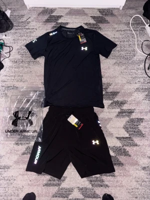 Svart träningsset från Under Armour - Snyggt svart träningsset från Under Armour med t-shirt och shorts. Båda plaggen har logga tryckt i vitt och reflekterande detaljer. Tillverkade i lätt och snabbtorkande material som passar perfekt för gym eller löpning. Shortsen har dragsko i midjan och tryck på benet.