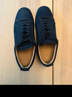 Louboutin  - Stilrena mörkblå sneakers i mocka med svarta skosnören och låg profil. Skorna har en klassisk design med rund tå och svart sula. Insidan är ljusbeige och materialet ger en soft och clean vibe. Perfekta för dig som gillar minimalistisk stil.