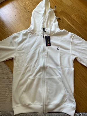 Vit hoodie från Polo Ralph Lauren - Vit hoodie med dragkedja från Polo Ralph Lauren. Klassisk design med huva, känguruficka och den ikoniska broderade loggan på bröstet. Tillverkad i mjukt material som känns skönt mot huden. Perfekt för en avslappnad och stilren look.