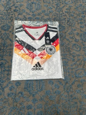 Tysklands matchtröja Adidas - Snygg vit matchtröja från Adidas med Tysklands landslagsmärke på bröstet. Tröjan har korta ärmar, V-ringning med röd kant och coolt mönster i svart, rött och gult över bröstet. Tillverkad i lätt och funktionellt material, perfekt för fotboll eller träning.