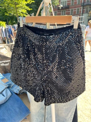 Svarta paljettshorts från Vero Moda - Glittriga svarta shorts från Vero Moda täckta med paljetter. De har en medelhög midja och kort passform, perfekta för att sticka ut. Materialet är syntet och shortsen har en mjuk insida för extra komfort.