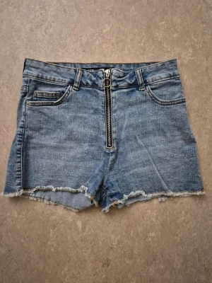 Jeansshorts - Säljer ett par blå jeansshorts med hög midja och råa kanter. Shortsen har en tydlig dragkedja framtill, fem fickor och bälteshällor. Perfekta för sommaren och ger en avslappnad vibe.