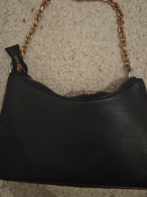 Svart handväska med guldkedja - Snygg svart handväska i konstläder med guldfärgad kedja som axelrem. Väskan har en enkel, stilren design och stängs med dragkedja upptill. Perfekt accessoar för att lyfta din outfit med en trendig touch.