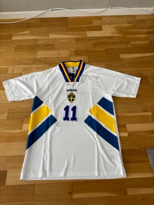Sverige bortaställ vm 94 Brolin#11 - Klassisk vit svensk landslagströja från Adidas med blå och gula detaljer, nummer 11 och Brolin på ryggen. Tröjan har V-ringad krage med blå och gula ränder samt broderad svensk landslagssköld på bröstet. Perfekt för fotbollsfans!