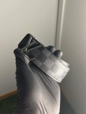 Rutigt svart bälte - Snyggt bälte från  med klassiskt rutigt mönster i svart och grått. Bältet har en stor -logga i silver som spänne och är gjort i skinn med en lyxig känsla. Perfekt accessoar för att lyfta din outfit.