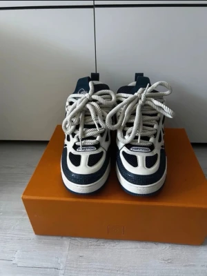 Louis Vuitton blåvita sneakers - Säljer ett par riktigt snygga Louis Vuitton sneakers i blått och vitt. Skorna har chunky sula, stora vita skosnören och ikoniska LV-blommor på sidan. Materialmix av läder och mesh ger en exklusiv känsla och cool kontrast. Perfekta för dig som vill sticka ut med lyxig streetstyle.