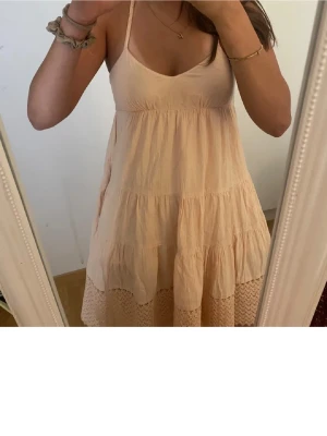 Ljus beige sommarklänning med flätade band - Supersöt ljusbeige klänning med flätade axelband och volangdetaljer. Klänningen är ärmlös, har v-ringad hals och faller i flera lager med spets längst ner. Perfekt för varma dagar och har en luftig, avslappnad passform.