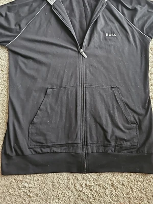 Svart ziptröja från BOSS Homewear - Mysig svart loungewear ziptröja från BOSS Homewear med vita detaljer och fickor framtill. Tröjan har dragkedja hela vägen, diskret BOSS-logga på bröstet och mjukt material som passar perfekt för chill hemma. Använd ett par gånger. Kommer att stryka den innan den skickas iväg. Använd max 10 gånger, inga skador. Size M