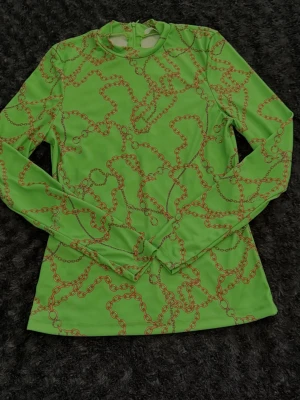 Grön långärmad topp med kedjemönster - Cool grön långärmad topp med orange kedjemönster. Toppen har en figurnära passform och dragkedja i nacken. Perfekt för dig som vill sticka ut med en unik och färgstark stil.