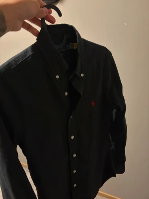 svart skjorta från Ralph Lauren - Klassisk svart skjorta från Ralph Lauren med knappkrage och vita knappar. Skjortan har lång ärm och den ikoniska röda logotypen broderad på bröstet. Tillverkad i mjuk bomull som känns skön mot huden. Den är aldrig knappt använd och har digitalt kvitto. Helt i nyskick