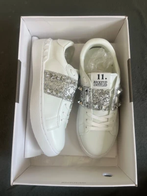 Valentino Rockstud Untitled sneakers - Snygga vita sneakers från Valentino med bredt band över foten dekorerat med silvriga nitar och glitterdetaljer. Modellen är låg och har klassisk rund tå samt vita skosnören. Märket och modellen står tryckt på plösen. Perfekta för dig som gillar statement-detaljer.