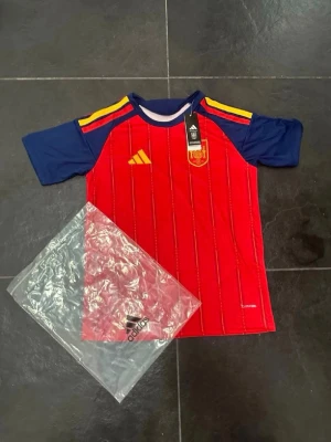 Spaniens matchtröja Adidas röd/blå - Säljer en officiell Spanien matchtröja från Adidas i rött med blå ärmar och gula detaljer. Tröjan har klubbmärke på bröstet, gula ränder på axlarna och är tillverkad i lätt funktionsmaterial. Perfekt för fotbollsträning eller att bära på matchdag.