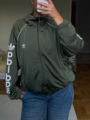 Grön Adidas ziptröja - Snygg grön vindjacka från Adidas med vit logga och text på ärmen. Jackan har dragkedja framtill och vita detaljer längs sömmarna. 