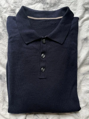 Mörkblå långärmad polotopp i Merinoull - Klassisk mörkblå långärmad polotopp från Charles Tyrwhitt med krage och tre knappar framtill. Tillverkad i mjukt merinoull som ger en clean och stilren look. Perfekt till jeans eller chinos för en enkel men snygg outfit.
