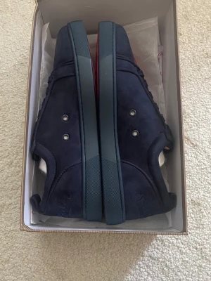 Christian Louboutin skor loubs mörkblå - Snygga mörkblå sneakers i mocka från Christian Louboutin. Skorna har lågt skaft, matchande mörkblå snörning och diskreta metalldetaljer på sidan. Klassisk röd detalj på sulan och stilren design som sticker ut.