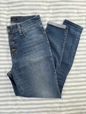 Blå high waist skinny jeans från Lucky Brand - Jeans från Lucky Brand i modellen High Rise Skinny Bridgette. Dessa jeans har en knäppning med flera knappar. De har fem fickor: två fram, en myntficka och två bakfickor. Jeansen har en mörkblå tvätt med slitna detaljer. De ser snygga ut både med och utan uppvikt benslut. På insidan av knapparna står det ”LUCKY YOU”. 