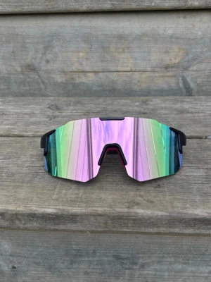 Pastellrosa Sportiga spegelglas solglasögon - Säljer ett par riktigt snygga solglasögon med spegelglas i regnbågsfärger och svart båge. De har en futuristisk, kantig form och passar perfekt för dig som vill sticka ut. Lätta och bekväma med cool vibe, perfekta för sommardagar.