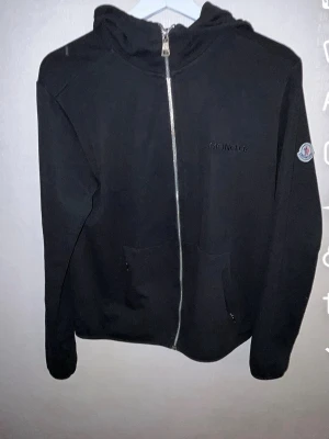 Svart hoodie med dragkedja från Moncler - Snygg svart hoodie från Moncler med dragkedja framtill och broderad Moncler-logga på bröstet. Klassisk huva och två fickor framtill. Materialet känns mjukt och bekvämt, perfekt för en avslappnad stil. Moncler-märke även på ärmen.