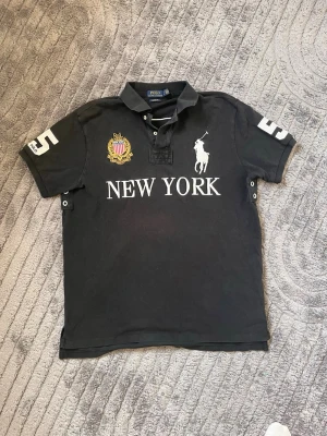 Svart pikétröja Polo Ralph Lauren - Svart pikétröja från Polo Ralph Lauren med broderad logga, stort tryck 'NEW YORK' på bröstet och siffran 5 på ärmarna. Klassisk krage med knappar och detaljer i vitt och guld. Perfekt för en sportig och stilren look. Det finns en liten märke under New York loggan som ser lite röd ut på bilden vet ej varför. Den är inte så där röd och syns inte mycket alls i verkligheten.