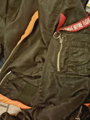 Svart bomberjacka Alpha Industries - Klassisk svart bomberjacka från Alpha Industries med ikonisk röd 'Remove Before Flight'-tag och orange foder. Jackan har dragkedja framtill och är tillverkad i slitstarkt nylonmaterial. Perfekt för dig som vill ha en tidlös och streetinspirerad look.