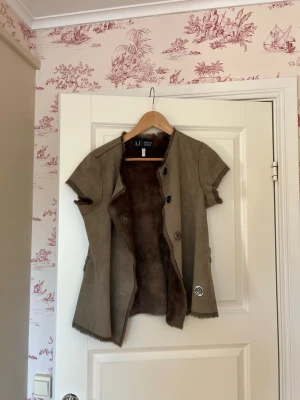 Brun pälsväst från AJ Mason - Säljer en brun pälsväst från AJ Mason med korta ärmar och knappar framtill. Västen har en mjuk pälsig utsida och slät insida, perfekt för att ge din outfit en lyxig touch. Klassisk rak passform och tidlös design.