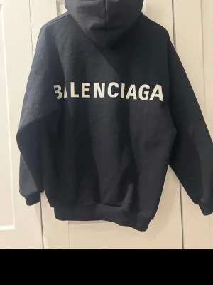 Svart hoodie från Balenciaga (oversized) - Snygg svart hoodie från Balenciaga med stor vit logga på ryggen. Klassisk oversized passform, huva och magficka framtill. Perfekt för dig som gillar streetwear och vill sticka ut med ett exklusivt plagg. Större i storleken än XXS. Pris kan diskuteras.