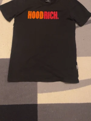 Svart HOODRICH t-shirt med tryck - Svart t-shirt från HOODRICH med färgglatt broderat tryck i orange och rött på bröstet. Klassisk passform med rund hals och korta ärmar. Perfekt för en streetwear-look.