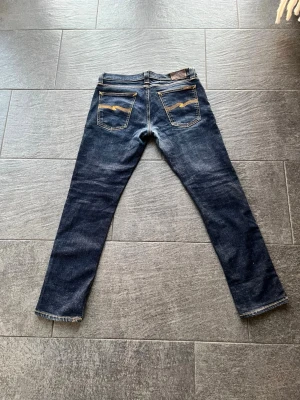 Mörkblå slim fit jeans från Nudie - Snygga mörkblå slim fit jeans från Nudie med klassiska orange sömmar och broderade bakfickor. Jeansen har 5 fickor och är tillverkade i denim med en smal passform som sitter tajt längs benen. Perfekta för en stilren och modern look.