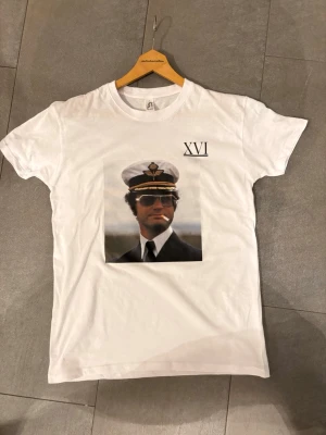 Vit t-shirt med marinmotiv och XVI-tryck - Vit t-shirt med tryck med kungen i marinuniform och texten 'XVI' på bröstet. Klassisk rund hals och korta ärmar. Perfekt för dig som gillar unika och grafiska plagg.   Tröjan finns i storlek S,Moch L 