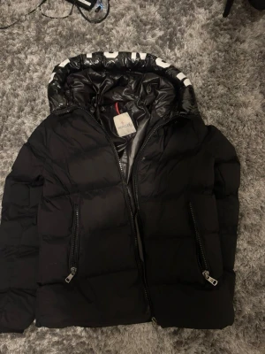 Svart pufferjacka från Moncler - Snygg svart pufferjacka från Moncler med stor huva och dragkedjor framtill. Jackan har en glansig insida på huvan och två praktiska fickor med dragkedja. Perfekt för kalla vinterdagar och riktigt trendig look.